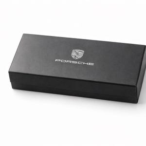 Op de afbeelding: Een zwarte rechthoekige doos met het Porsche-logo en de naam in zilver. De doos staat op een witte achtergrond. Het Porsche-embleem is een schild met het woord "Porsche" erboven.