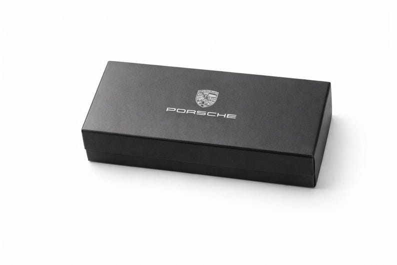 Op de afbeelding: Een zwarte rechthoekige doos met het Porsche-logo en de naam in zilver. De doos staat op een witte achtergrond. Het Porsche-embleem is een schild met het woord "Porsche" erboven.