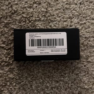 Op de afbeelding: Een zwarte rechthoekige doos met een wit etiket. Het etiket bevat de tekst "WAP0500310NWSA" en "Key fob Black". De doos staat op een getextureerd, bruin oppervlak. Gemaakt in Duitsland.