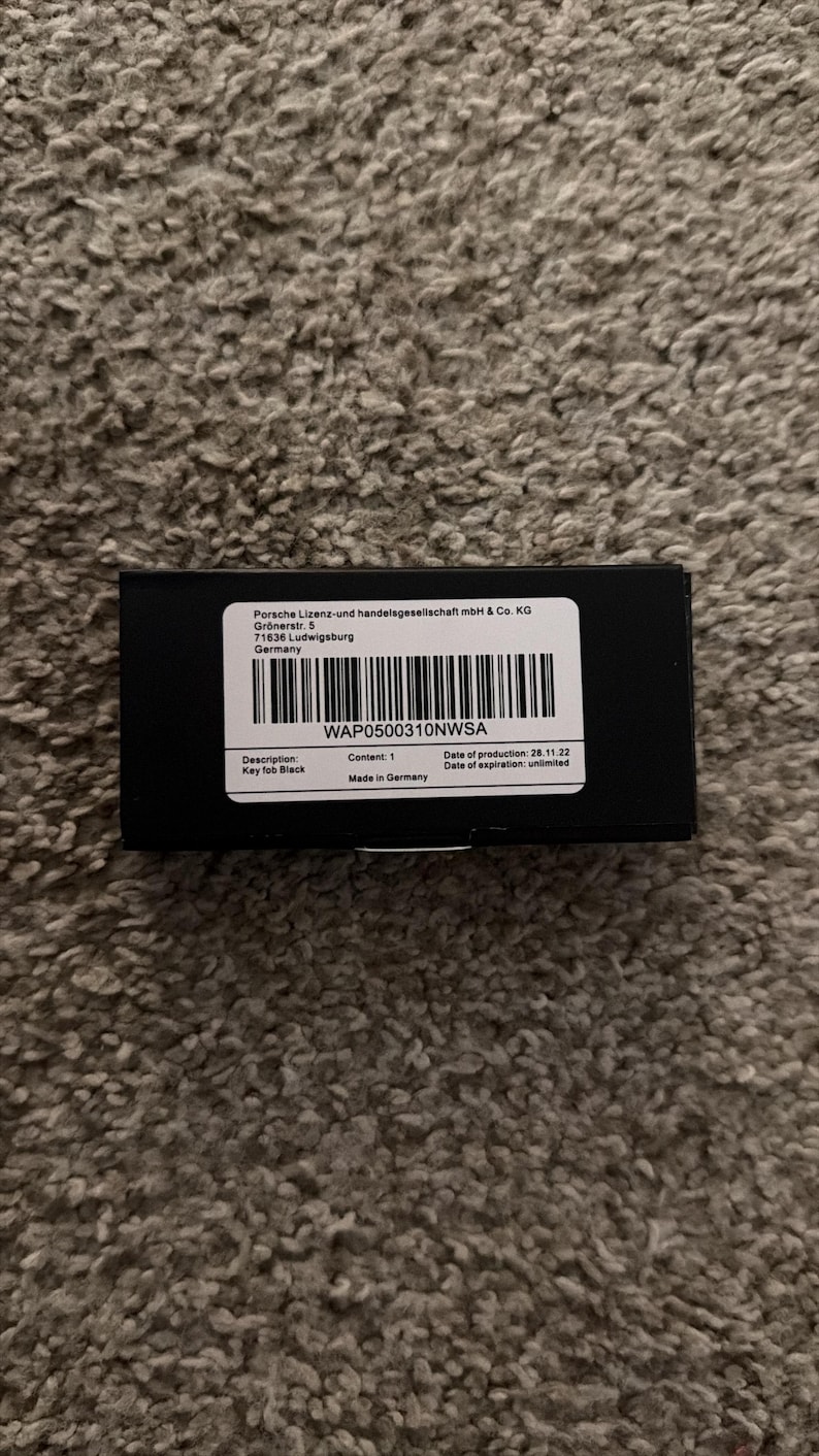 Op de afbeelding: Een zwarte rechthoekige doos met een wit etiket. Het etiket bevat de tekst "WAP0500310NWSA" en "Key fob Black". De doos staat op een getextureerd, bruin oppervlak. Gemaakt in Duitsland.