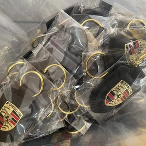 Op de afbeelding: Een verzameling zwarte sleutelhangers met goudkleurige sleutelringen, elk met het Porsche-embleem in rood, goud en zwart. De sleutelhangers zijn afzonderlijk verpakt in doorzichtig plastic.