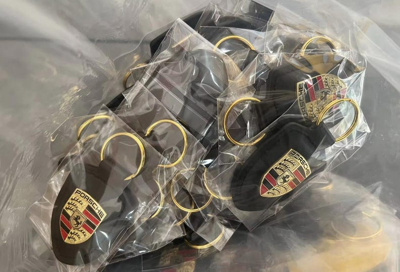 Op de afbeelding: Een verzameling zwarte sleutelhangers met goudkleurige sleutelringen, elk met het Porsche-embleem in rood, goud en zwart. De sleutelhangers zijn afzonderlijk verpakt in doorzichtig plastic.
