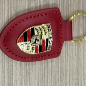 Op de afbeelding: Rode leren sleutelhanger met een goudkleurige ring. De sleutelhanger heeft een goud, zwart en rood embleem met het woord "PORSCHE" in gouden letters. De sleutelhanger is gestikt met rode draad.