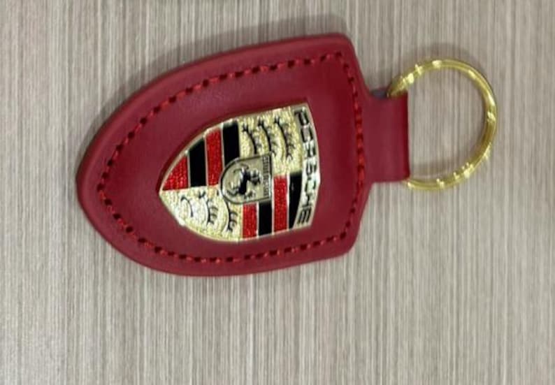 Op de afbeelding: Rode leren sleutelhanger met een goudkleurige ring. De sleutelhanger heeft een goud, zwart en rood embleem met het woord "PORSCHE" in gouden letters. De sleutelhanger is gestikt met rode draad.