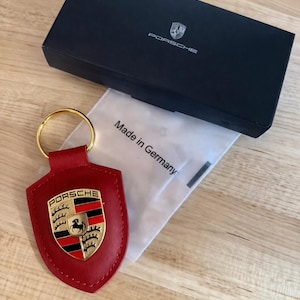 May include: Red leather keychain with a gold ring and a Porsche emblem. The keychain is shield-shaped and features the Porsche logo in gold, black, and red. A black box with the Porsche logo and the text "Made in Germany" is also visible.