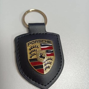 Op de afbeelding: Zwarte leren sleutelhanger met een gouden ring en een Porsche-embleem. Het embleem toont het Porsche-logo met het woord "PORSCHE" bovenaan en "STUTTGART" onderaan. Het embleem is goud, rood en zwart.