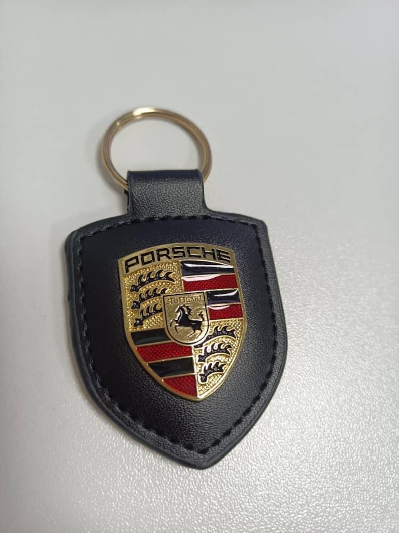 Op de afbeelding: Zwarte leren sleutelhanger met een gouden ring en een Porsche-embleem. Het embleem toont het Porsche-logo met het woord "PORSCHE" bovenaan en "STUTTGART" onderaan. Het embleem is goud, rood en zwart.