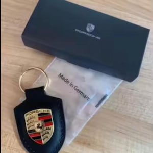 Op de afbeelding: Een zwarte leren Porsche sleutelhanger met een gouden sleutelring, met het Porsche embleem in rood, goud en zwart. De sleutelhanger ligt naast een zwarte doos met het Porsche logo en een zakje met "Made in Germany".