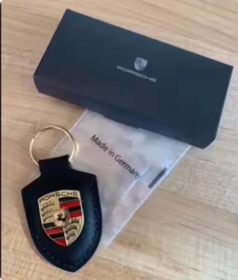 Op de afbeelding: Een zwarte leren Porsche sleutelhanger met een gouden sleutelring, met het Porsche embleem in rood, goud en zwart. De sleutelhanger ligt naast een zwarte doos met het Porsche logo en een zakje met "Made in Germany".