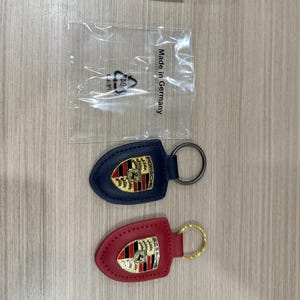 Op de afbeelding: Twee leren sleutelhangers met het Porsche-logo. E&eacute;n is marineblauw met een zilverkleurige ring, de andere is rood met een goudkleurige ring. Ze liggen op een licht houtnerfoppervlak. Een plastic zak met tekst staat op de achtergrond.