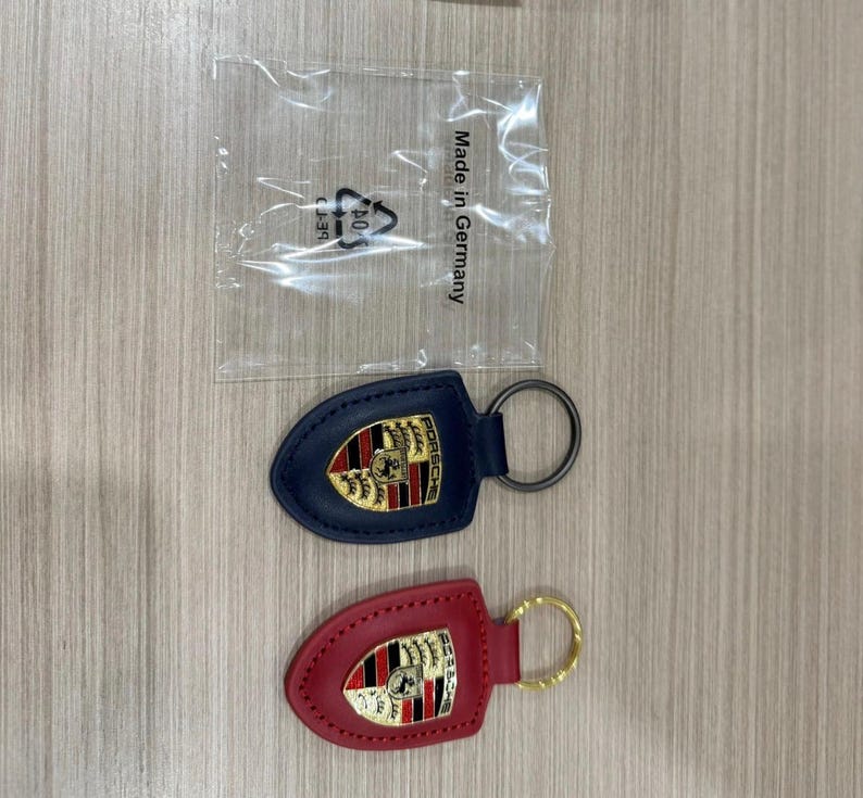 Op de afbeelding: Twee leren sleutelhangers met het Porsche-logo. E&eacute;n is marineblauw met een zilverkleurige ring, de andere is rood met een goudkleurige ring. Ze liggen op een licht houtnerfoppervlak. Een plastic zak met tekst staat op de achtergrond.