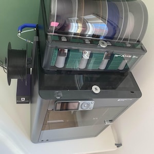 Może przedstawiać: Czarno-szara drukarka 3D z przezroczystym blatem, wyświetlająca wiele szpul filamentu w różnych kolorach. Drukarka posiada wyświetlacz cyfrowy i boczny uchwyt na szpulę. Urządzenie stoi na białym stole.