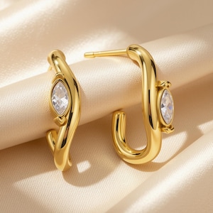 May include: A pair of gold-toned hoop earrings featuring a smooth, curved metal design. Each earring is accented with a single marquise-cut clear gemstone set into the center of the hoop, complemented by a post-back closure mechanism.