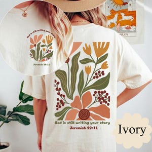 Puede incluir: Camiseta de color marfil con un diseño floral en tonos naranja, verde y amarillo. El texto "God is still writing your story Jeremiah 29:11" está impreso en la parte delantera y trasera. La camiseta tiene cuello redondo y mangas cortas.