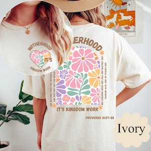 Puede incluir: Una camiseta color marfil con un gráfico frontal de un corazón con flores y el texto MOTHERHOOD IT'S KINGDOM WORK. La parte trasera muestra un arco grande lleno de flores coloridas, el texto MOTHERHOOD, IT'S KINGDOM WORK y PROVERBS 31:27-28.