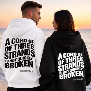Puede incluir: Dos sudaderas con capucha, una blanca y otra negra, con la frase "A CORD OF THREE STRANDS IS NOT QUICKLY BROKEN" y la cita bíblica Eclesiastés 4:12. Las sudaderas las llevan dos personas, con un atardecer de fondo.