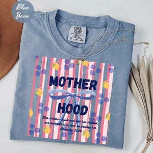 Puede incluir: Una camiseta color azul vaquero con un gráfico de rayas verticales rosas y blancas, bayas azules, limones amarillos y un lazo azul. El texto dice MOTHER HOOD y Her children arise and call her blessed; her husband also, and he praises her Proverbs 31:28.