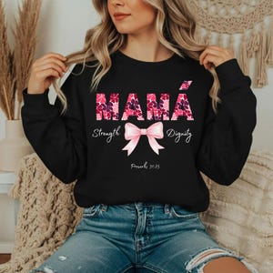 Puede incluir: Sudadera negra con la palabra "MAMÁ" en letras florales rosas. Debajo, un lazo rosa y las palabras "Fuerza" y "Dignidad". El texto "Proverbios 31:25" está impreso debajo del lazo.