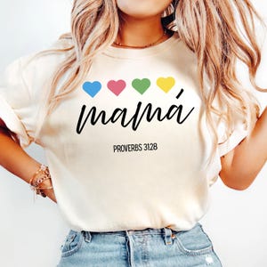 Puede incluir: Camiseta color crema con la palabra "mamá" en escritura negra, acentuada por cuatro corazones coloridos. El texto "PROVERBIOS 31:28" está impreso debajo. La camiseta es de estilo casual.