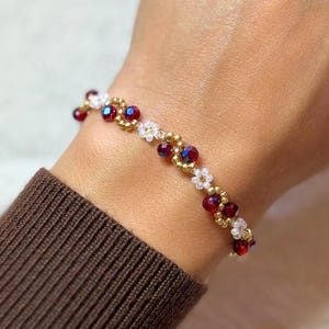 May include: A beaded bracelet featuring red and white beads with gold accents. The bracelet is adorned with small, flower-shaped beads and is worn on a wrist with a brown sweater sleeve.