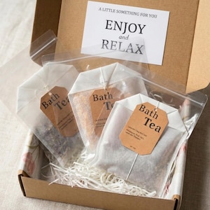 May include: A brown cardboard gift box containing three white fabric bath tea bags, each with a brown paper tag labelled Bath Tea. A white card inside the box lid features the text A LITTLE SOMETHING FOR YOU ENJOY and RELAX in black font.