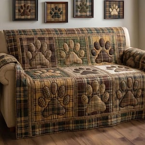 May include: A plaid sofa cover with large paw print appliques in shades of brown, green, and tan. The cover is draped over a beige sofa. Framed paw print art hangs above the sofa.