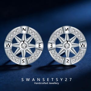 Puede incluir: Un par de pendientes plateados con forma de brújula. Cada pendiente presenta un diseño de rosa de los vientos con los puntos cardinales. El anillo exterior está incrustado con pequeñas gemas transparentes. Los pendientes están sobre un fondo azul oscuro.