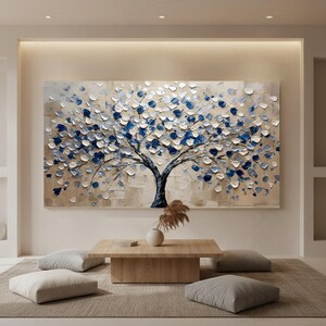 Op de afbeelding: Groot abstract schilderij van een boom met blauwe en witte getextureerde bladeren tegen een beige achtergrond. De boomstam is donkerblauw. Het schilderij hangt boven een houten salontafel met een vaas en decoratieve kussens.