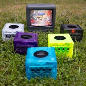 May include: A collection of colorful, translucent video game consoles arranged on grass, surrounding a gray CRT television displaying a video game. The consoles are in clear, purple, blue, green, and black. The TV shows the Sonic Adventure 2 title screen.