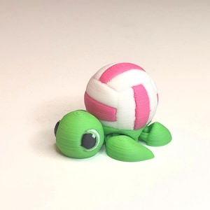 May include: A 3D-printed turtle figurine with a volleyball-shaped shell. The shell is white with pink stripes, and the turtle's head and flippers are green. The eyes are black. The figurine is on a white surface.