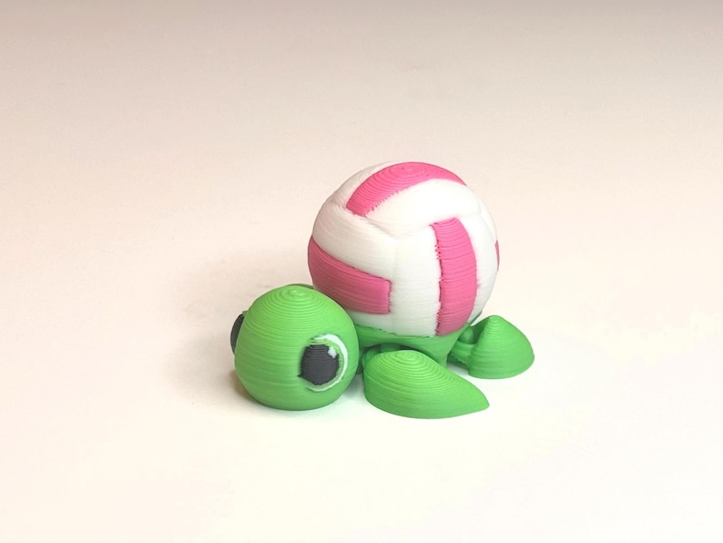 May include: A 3D-printed turtle figurine with a volleyball-shaped shell. The shell is white with pink stripes, and the turtle's head and flippers are green. The eyes are black. The figurine is on a white surface.