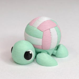 May include: A whimsical, mint green turtle figurine with a volleyball shell. The turtle's head has large, black-pupiled eyes. The volleyball is white, pink, and mint green. The figurine is a unique decorative item.