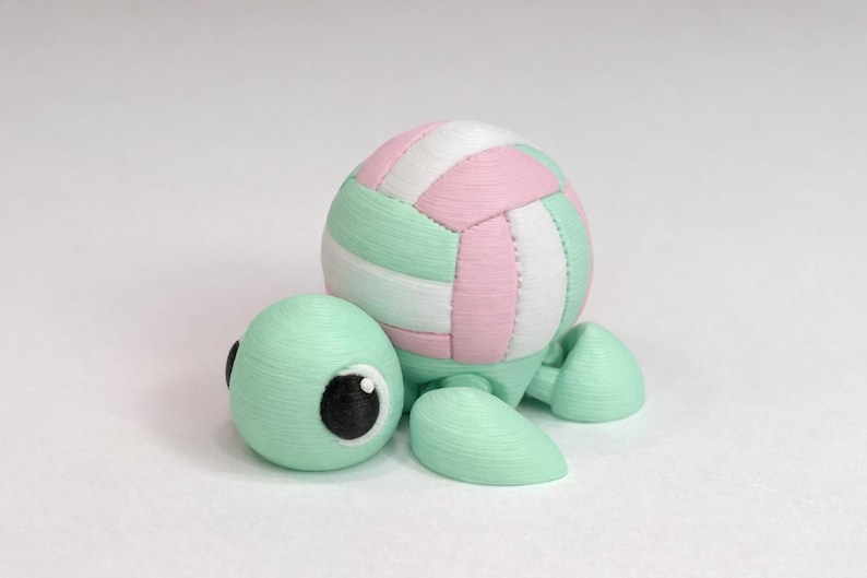 May include: A whimsical, mint green turtle figurine with a volleyball shell. The turtle's head has large, black-pupiled eyes. The volleyball is white, pink, and mint green. The figurine is a unique decorative item.