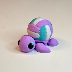 May include: A 3D-printed turtle figurine with a volleyball-shaped shell. The turtle is primarily lavender, with a multi-colored shell featuring teal, white, and lavender stripes. The turtle has black eyes and is set against a white background.