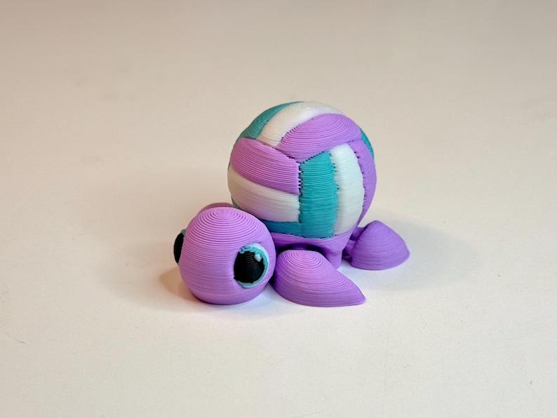 May include: A 3D-printed turtle figurine with a volleyball-shaped shell. The turtle is primarily lavender, with a multi-colored shell featuring teal, white, and lavender stripes. The turtle has black eyes and is set against a white background.