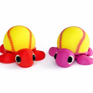 May include: Two whimsical turtle figurines. Each turtle has a yellow softball for a shell, with red and pink bodies. The turtles have black eyes and are set against a white background.