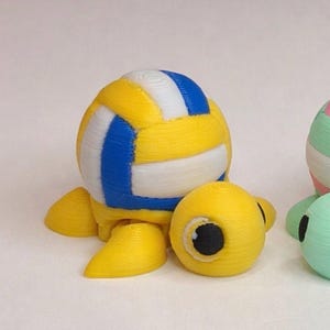 May include: A 3D-printed yellow turtle figurine with a volleyball shell. The volleyball has blue and white stripes. The turtle has yellow flippers and a yellow head with a black eye.