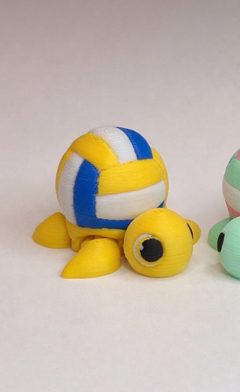 May include: A 3D-printed yellow turtle figurine with a volleyball shell. The volleyball has blue and white stripes. The turtle has yellow flippers and a yellow head with a black eye.