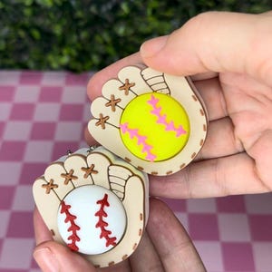 May include: Two baseball glove charms, one featuring a bright yellow softball with pink stitching and the other a white baseball with red stitching. Both gloves are light tan with brown lacing details and a rounded, flat-backed design.