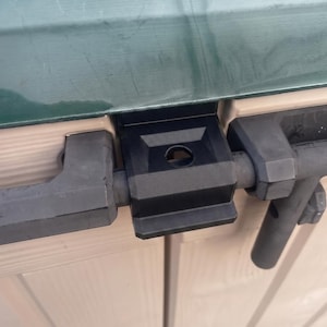 Peut inclure: Gros plan sur un loquet de cabanon noir et gris. Le loquet est en plastique et en métal, avec un mécanisme de boulon coulissant. L'extérieur de l'abri est beige clair et vert.