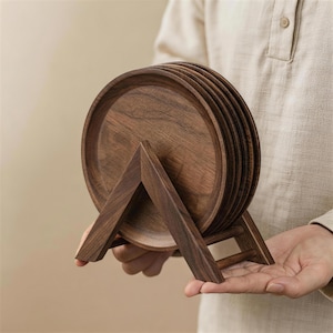 May include: A set of six round, dark brown wooden coasters on a matching wooden stand. The coasters have a slightly raised edge and are held in place by a triangular stand. The stand has a simple, minimalist design.