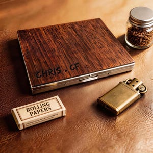 May include: A wooden box with the engraved text "CHRIS . CF" sits on a brown surface, next to a small box of rolling papers, a gold-colored lighter, and a small glass jar with a silver lid.