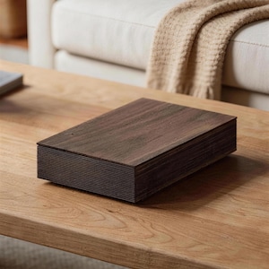May include: A dark brown wooden box, rectangular in shape, composed of many thin layers, possibly for storage or decoration. It rests on a light brown wooden table, with a neutral-toned sofa and blanket visible in the background.