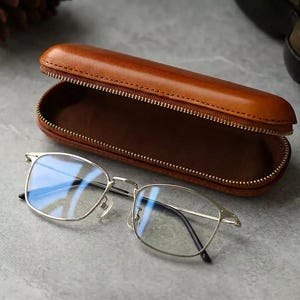 Könnte beinhalten: Eine silberfarbene Metallbrille mit klaren Gläsern vor einem offenen, länglichen, hellbraunen Lederetui mit goldenem Reißverschluss und dunkelbraunem Innenfutter, das zur schützenden Aufbewahrung der Brille dient.