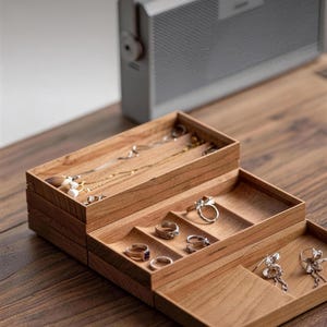 Puede incluir: Un organizador de joyas de madera de varios niveles con un acabado de grano de madera marrón claro natural. La bandeja superior contiene collares, la bandeja central presenta secciones divididas para anillos y la inferior muestra pendientes colgantes.