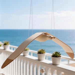 May include: A wooden seagull mobile with outstretched wings, suspended from a wooden dowel. The seagull has a natural wood finish with brown accents on the wingtips. The background features a white railing, small potted plants, and the ocean.