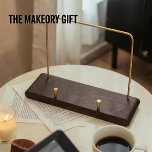 May include: A dark brown wooden jewelry display stand with two gold pegs and a gold metal bar. The text "THE MAKEOORY GIFT" is at the top. A lit candle, a cup of coffee, and papers are on the table.