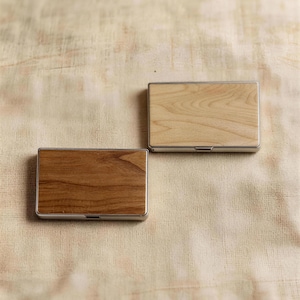 Puede incluir: Dos tarjeteros rectangulares con acabado de vetas de madera. Uno es de color madera clara y el otro de color madera más oscuro. Ambos tienen un marco de metal plateado. Los tarjeteros se muestran sobre una superficie de tela beige.