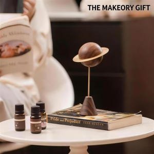 May include: A wooden planet sculpture with a gold ring and stand, displayed on a table with a book titled "Pride and Prejudice" and essential oil bottles. The text "THE MAKEOY GIFT" is visible.