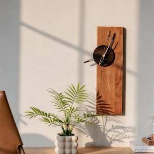 May include: A wooden wall clock with a dark circular face and black hands. The clock is mounted on a light-coloured wall. A small potted plant with green leaves sits on a wooden table in front of the clock.